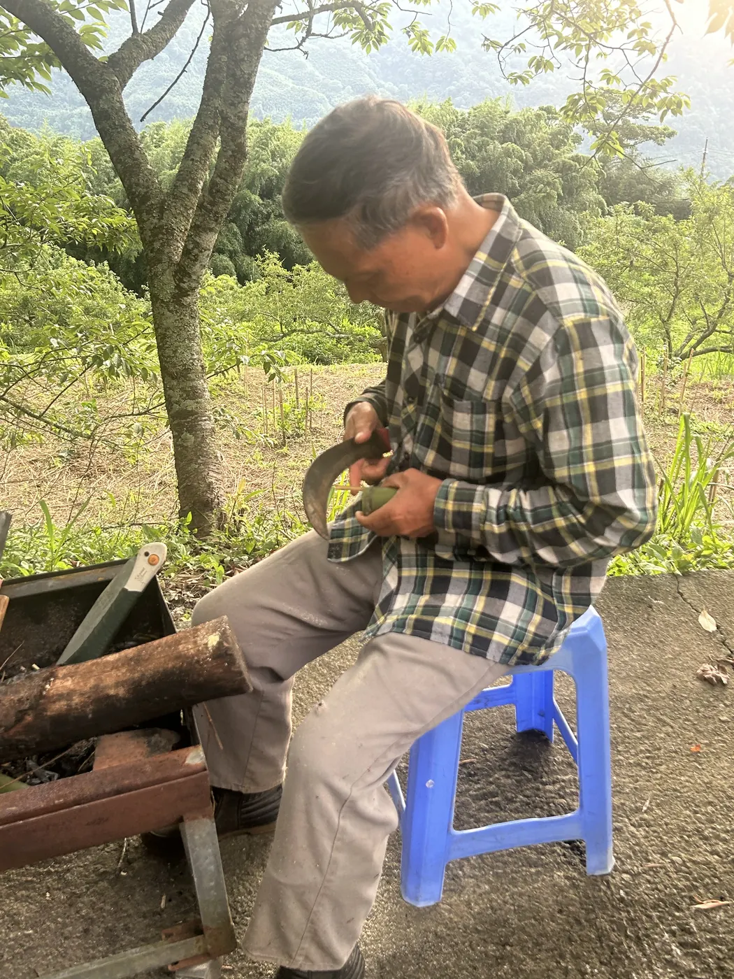 Yaba 坐在果園邊，以山刀削製竹杯，背後是果樹與遠山